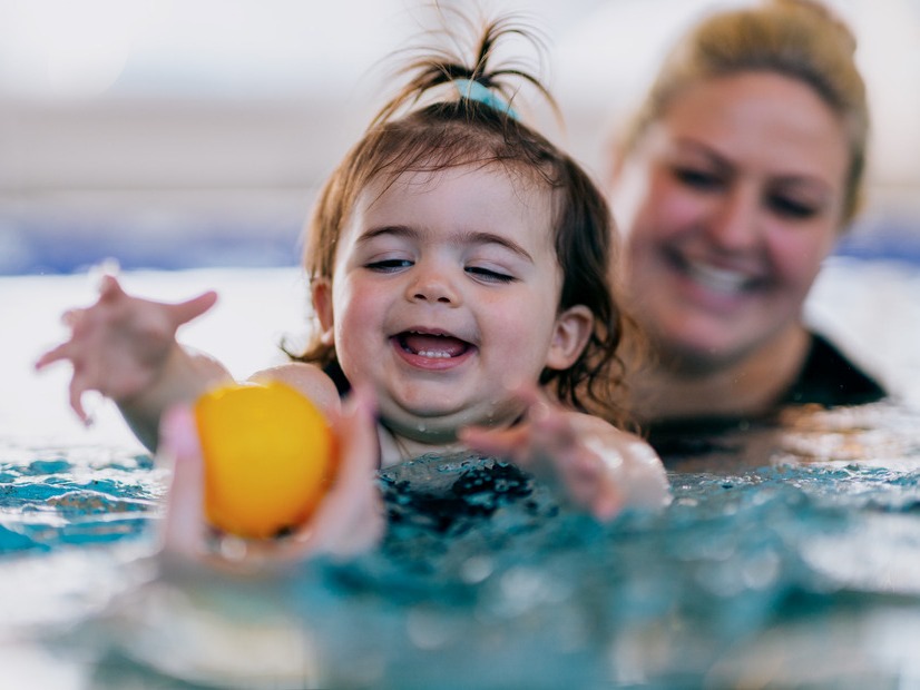 Toddler reaching for a ball in the pool, supported by a Kingswim teacher