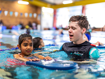 Girl learning to swim on her front with a float, supported by a swim teacher